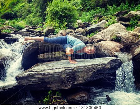 Vintage retro effect hipster style image of sporty fit woman doing yoga asana Eka Pada Koundinyasana 1 at tropical waterfall