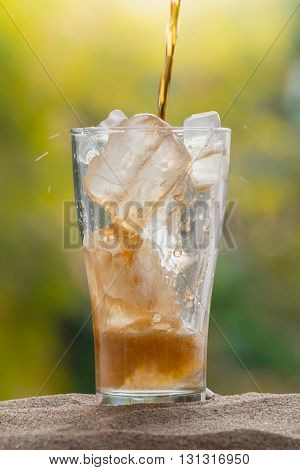 Cola water pouring into glass with ice cubes on sand nature background