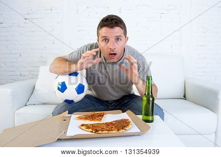 young man alone holding ball in stress watching football game on television sitting at home living room sofa couch with pizza box and beer bottle enjoying the match looking excited and shocked