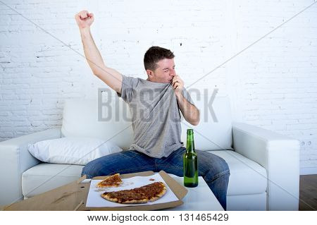 young man watching football game on television celebrating goal crazy happy on sofa couch at home with ball beer bottle and pizza looking excited and cheerfull kissing his team shield