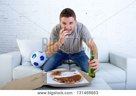 young man watching football game on television nervous and excited suffering stress biting fingernail on sofa couch at home with ball holding beer bottle eating pizza looking crazy anxious