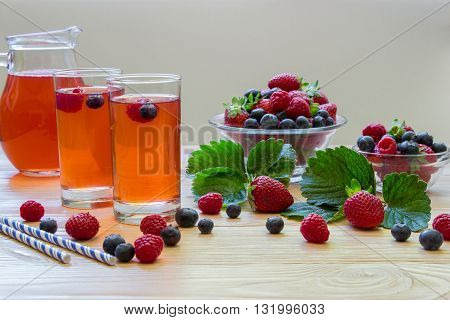 Left two glasses of berries compote near 2 bowls with berries carafe compote on a light wooden background scattered berries and some empty space. Fresh berries compote and empty space. Horizontal.