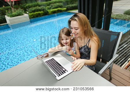 Happy Parent Mother And Daughter Using Laptop On Background Of Swimming Pool During Sunny Day. Conce