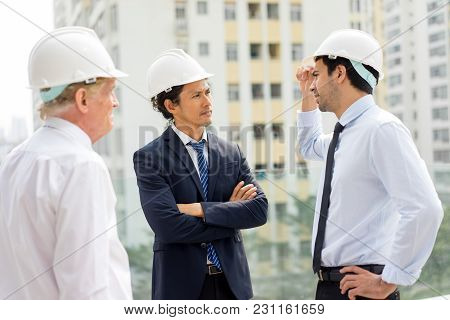 Strict Man In Helmet And Suit With Folded Arms Listening To Two Technical Experts. Two Engineers Rep