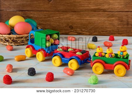 Color Easter Sweets In Toy Track And Trailers Next To Color Easter Eggs And Candy On Wooden Backgrou
