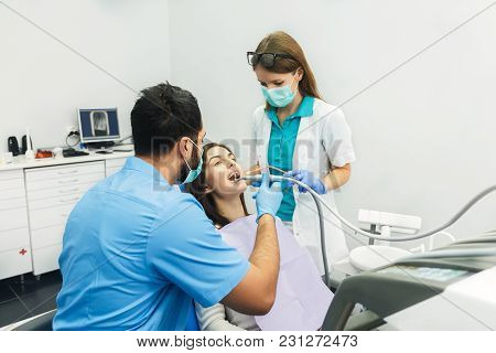 Smiling Beautiful Patient Sitting In Chair With Dental Purple Bib, Male Dentist, In Blue Uniform And