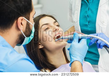 Smiling Beautiful Patient Sitting In Chair, Male Dentist, In Blue Uniform And Gloves, Holding Explor