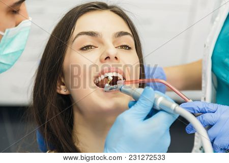 Smiling Beautiful Patient Sitting In Chair, Male Dentist, In Blue Uniform And Gloves, Holding Explor