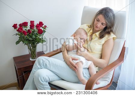Young Mother Holding Her Baby, Rocking Him To Sleep, Sitting In Armchair