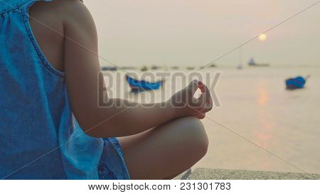 Little Cute Girl Meditates In Turkish Pose At Seafront With The Sea And Fishing Boats On The Backgro