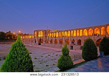 The Khaju Bridge In Evening Lights And Dried Up Zayandeh River Are Seen Through The Young Thuja Tree