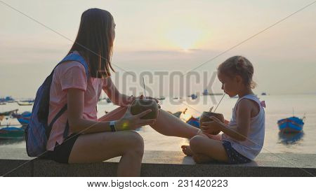 Travelers Mother And Little Daughter Sits At Seafront And Drinks Coconats In Trip. Happy Family Is E