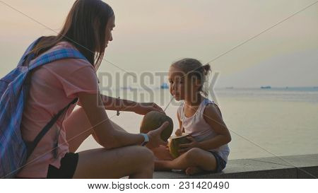 Travelers Mother And Little Daughter Sits At Seafront And Drinks Coconats In Trip. Mom Shares Her Co