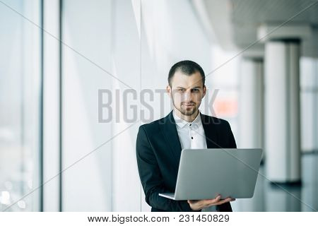 Businessman Staring Intently At The Camera As He Stands In A Bright Airy Office Holding An Open Lapt