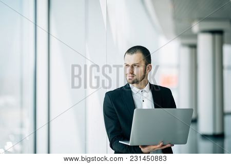 Businessman Staring Intently At The Camera As He Stands In A Bright Airy Office Holding An Open Lapt