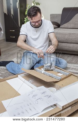 The Man Kneels On The Carpet Above The Cardboard Box With Elements Of Furniture For Self-assembly An