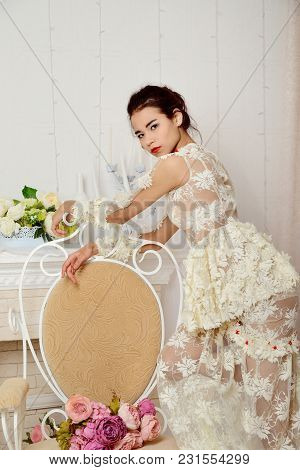 Asian Girl In Beautiful Dress Posing In The Studio Beside The Chair On Which Lies A Bouquet Of Flowe