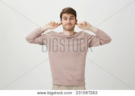 Indoor Portrait Of Confused Smiling Caucasian Man Covering Ears With Forefingers, Standing With Awkw