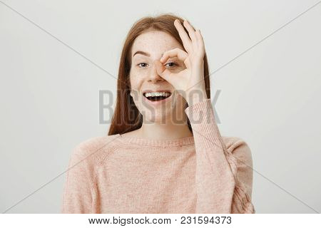 Studio Portrait Of Positive Cute Redhead Woman Showing Okay Sign Over Eye And Looking Through It At 