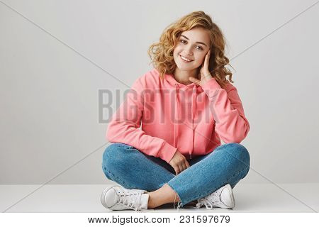 Studio Portrait Of Cute Female Freelancer With Curly Hair Sitting On Floor With Crossed Lefs Leaning