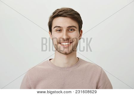 Confident Attractive Caucasian Guy In Beige Pullover Smiling Broadly While Standing Against Gray Bac
