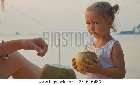 Travelers Mother And Little Daughter Sits At Seafront And Drinks Coconats In Trip. Portrait Of Cute 