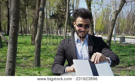 Attractive Person Sits By Table, Smiling With Dimples On Cheeksand Uses Laptop. Guy Has Short Dark H