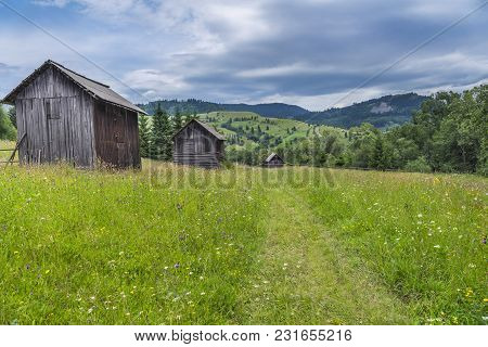 Rustic Landscape With Old Barns In A Row, On A Field With Green Grass And Colorful Wildflowers, Cros