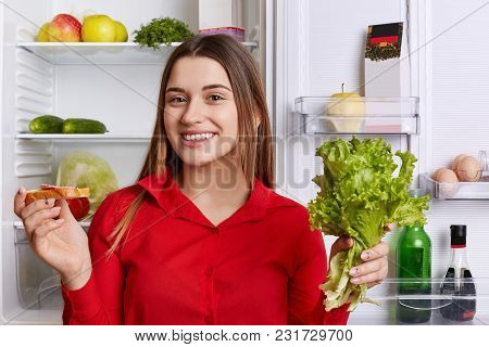 Adorable Young Woman With Happy Expression, Dressed In Red Blouse, Holds Lettuce And Sandwhich While