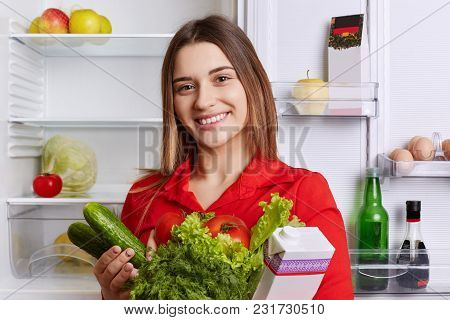 Satisfied Woman Holds Fresh Vegetables And Milk, Going To Put Them In Refridgerator, Has Broad Smile