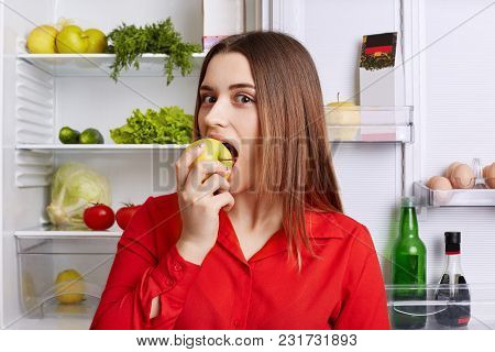 Good Looking Female Model Bited Delicious Apple, Stands Near Opened Fridge Full Of Healthy Products,