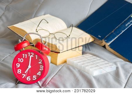 Red Alarm Clock With Open Book On Strack And Pills Box On Bed