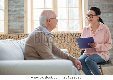 Duties Of Caregiver. Appealing Glad Caregiver Using Clipboard While Wearing Glasses And Talking To S