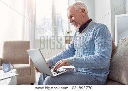 Mood-boosting Chat. Upbeat Senior Man Sitting On The Couch And Typing An Email On The Laptop While S