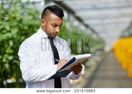 Young serious engineer in whitecoat making notes while working in modern hothouse