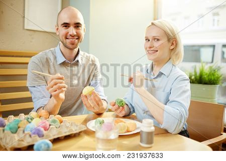 Young cheerful man and woman looking at camera while painting eggs for Easter