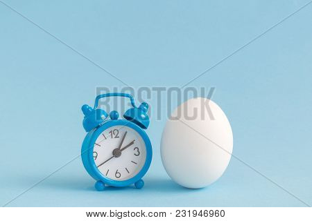 Small Alarm Clock And White Egg On Pastel Blue Background. Eastertime Minimal Concept.