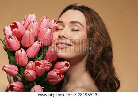 Beauty portrait of a pretty young topless woman with brown curly hair holding floers with eyes closed isolated over beige background