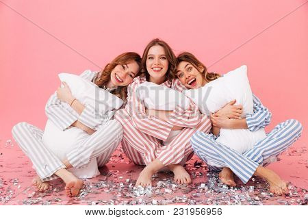 Photo of happy women 20s wearing leisure clothings sitting barefoot on floor with legs crossed and hugging pillows during girlish sleepover isolated over pink background