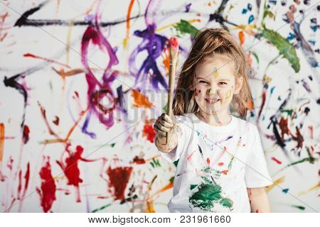 Cute child with smuges of colorful paint showing a paint brush. Little artist.