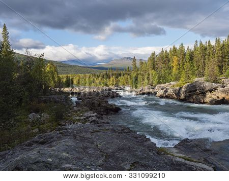 Beautiful Northern Landscape With Wild Glacier River Kamajokk, Boulders And Spruce Tree Forest In Kv