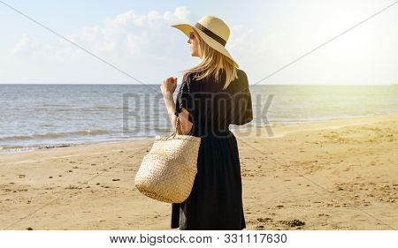 Side View Of Young  Caucasian Woman With Black Dress  ,  Straw Hat And Basket Waliking  On Beach   A