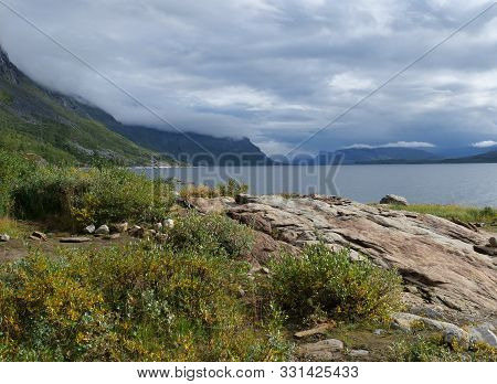 View On Akkajaure Lake With Green Mountains, Rock Boulders, Bushes, Birch Tree And Dramatic Clouds