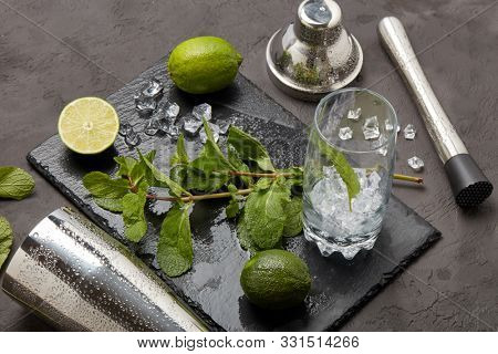 Set of bar tools: cocktail shaker, muddler and glass with ice, lime and mint  on dark stone  table. Top view. Bartender's tool. Mojito cocktail making.