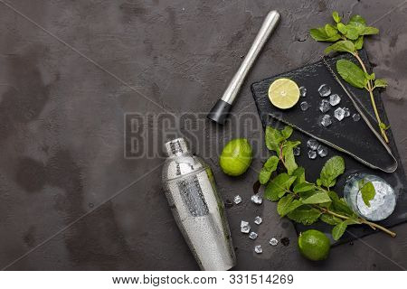 Set of bar tools: cocktail shaker, muddler, ice tongs and glass with ice, lime and mint  on dark stone  table. Top view with copy space. Bartender's tool. Mojito cocktail making.