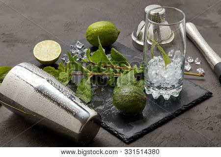 Set of bar tools: cocktail shaker, muddler and glass with ice, lime and mint  on dark stone  table. Top view. Bartender's tool. Mojito cocktail making.