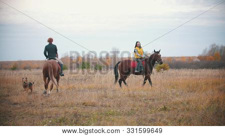 Two Women Riding Horses In The Autumn Field - A Dog Walking Near Them