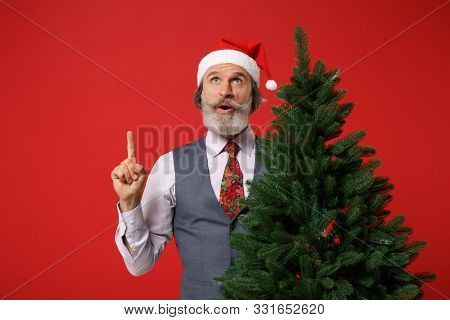 Amazed Elderly Gray-haired Mustache Bearded Man In Santa Hat Shirt, Vest Tie Isolated On Red Backgro