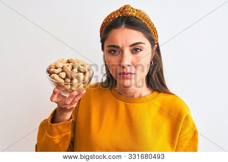 Young beautiful woman holding bowl with peanuts standing over isolated white background with a confident expression on smart face thinking serious