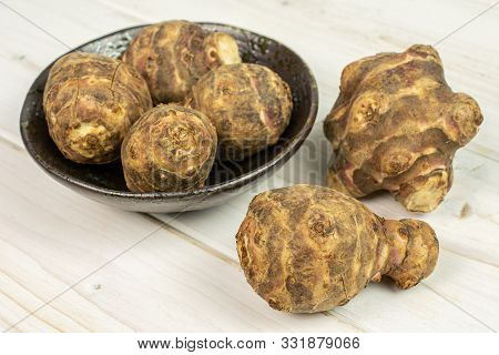 Group Of Six Whole Fresh Brown Jerusalem Artichoke In Dark Ceramic Bowl On White Wood